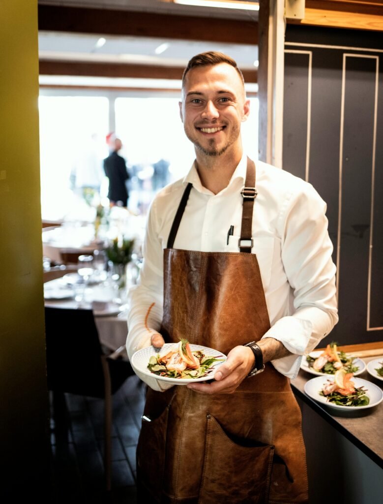 A smiling waiter in a white shirt and brown apron holding a delicious dish, indoors in a Danish restaurant.